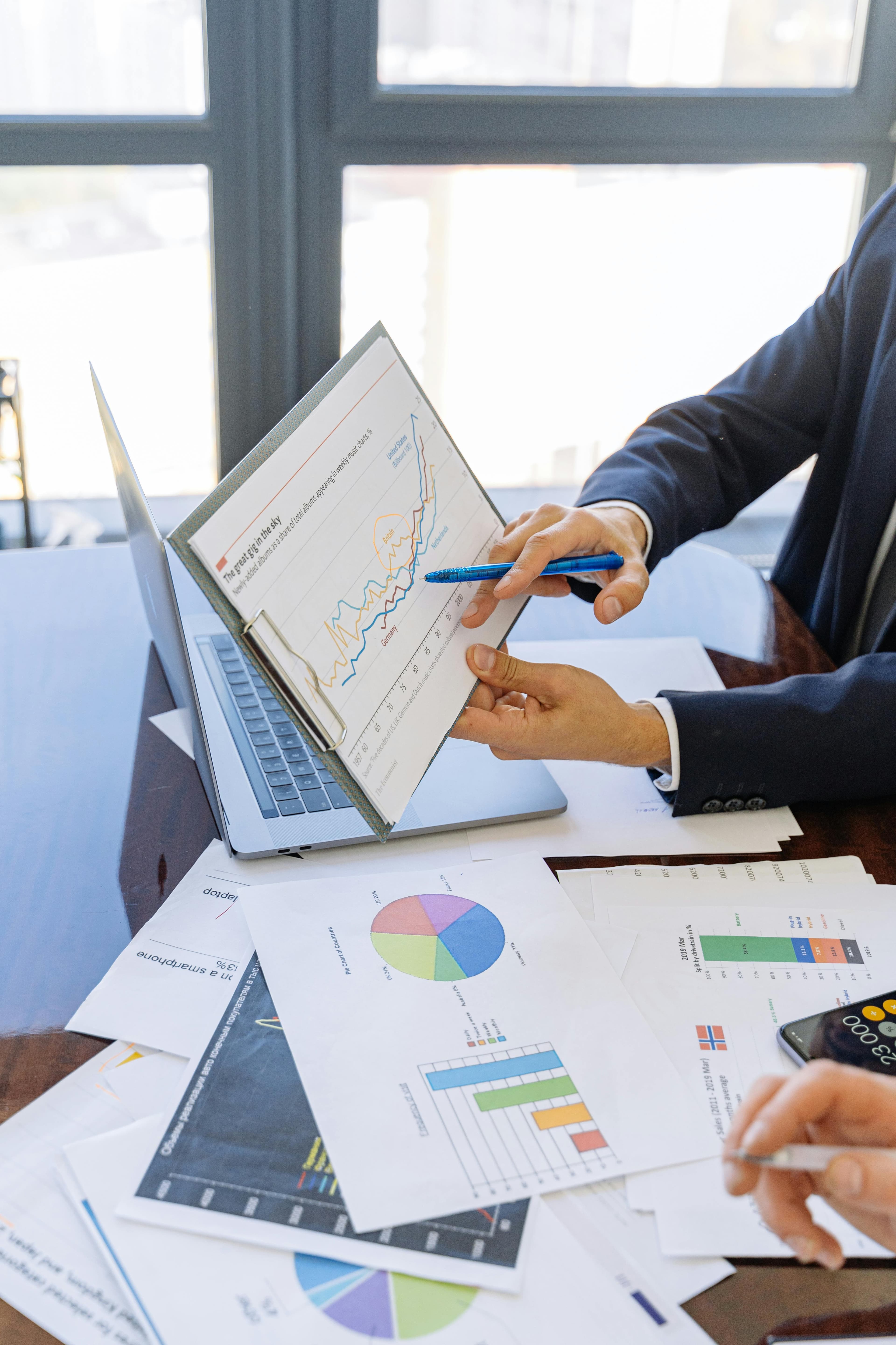 Businessman pointing at a line graph on a clipboard over a desk with financial charts.